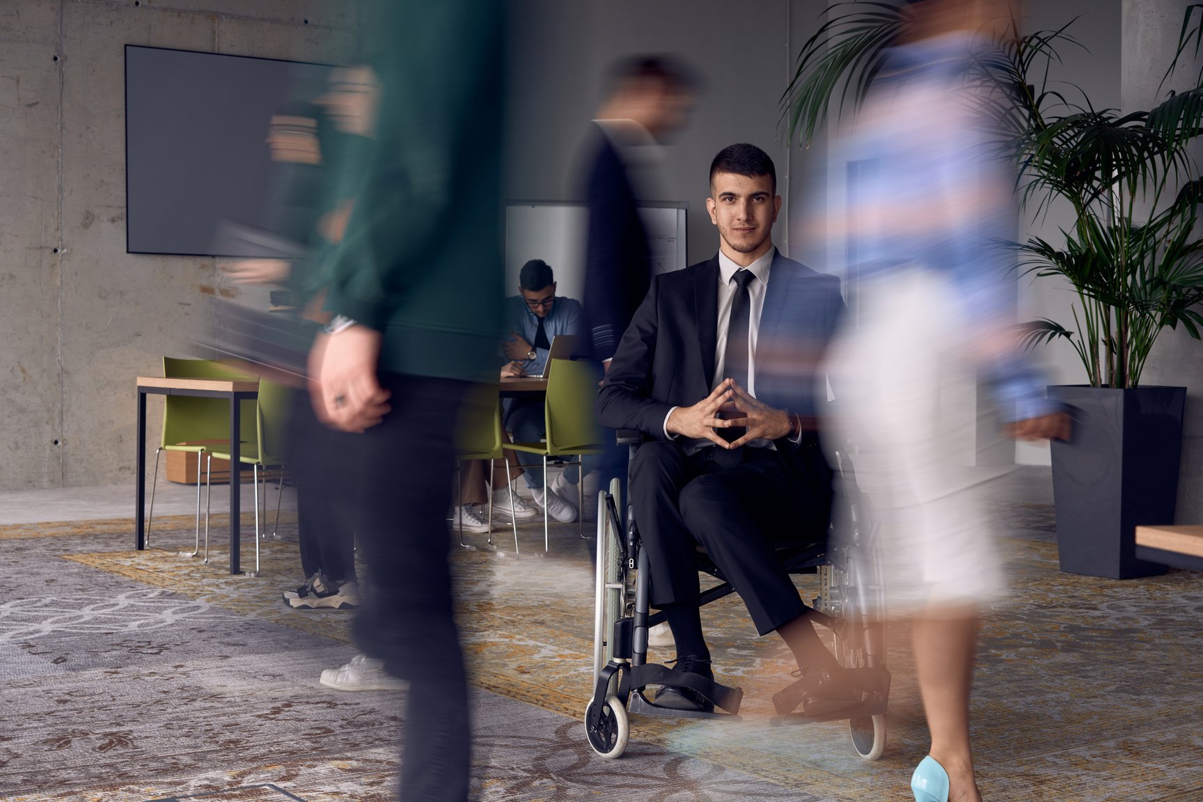 A businessman in a wheelchair in a modern office, surrounded by his colleagues who are portrayed with blurred movements, symbolizing their support and solidarity as they navigate the workspace together.